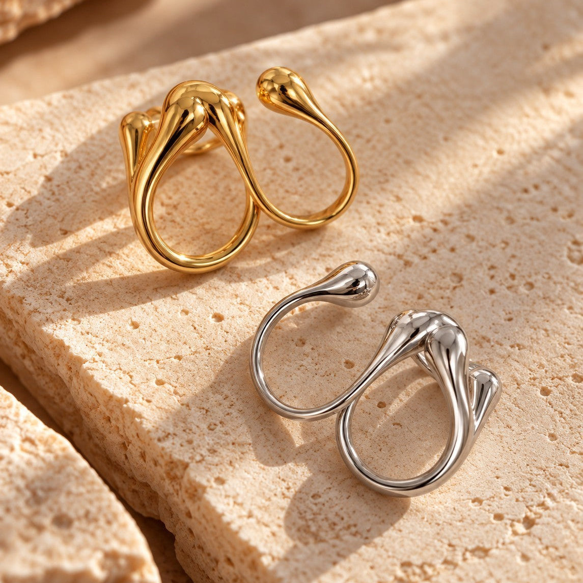 Reneax and Company sculptural double finger rings in polished gold and silver tones on a textured stone surface.